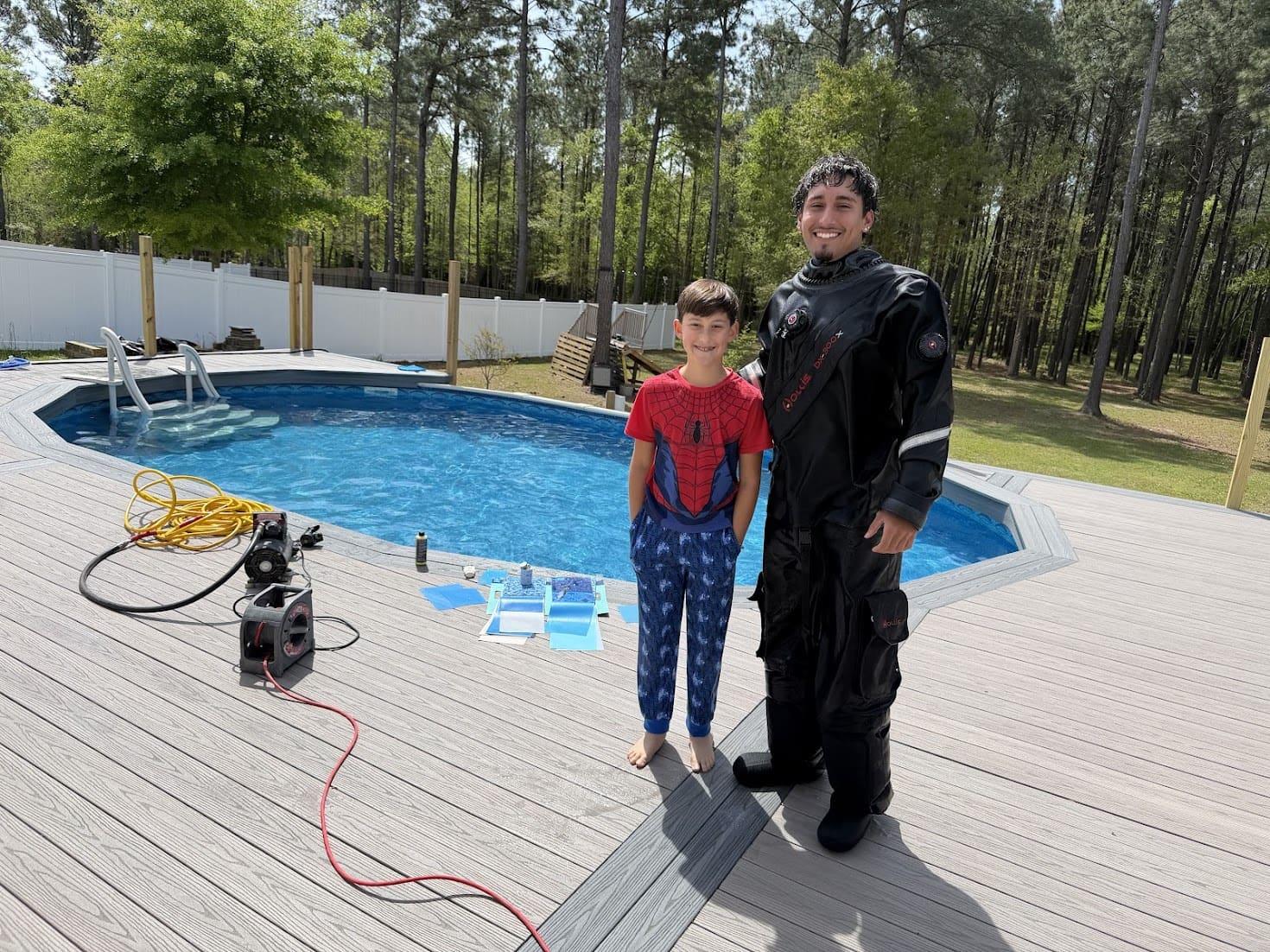 Leak-detection technician in a drysuit standing with a young boy beside a residential above-ground pool, with liner patch material and equipment on the deck.